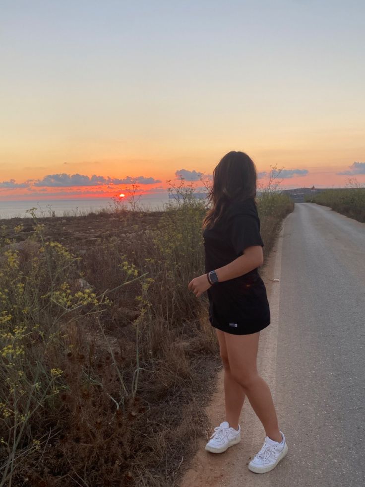 a woman standing on the side of a road next to grass and bushes at sunset