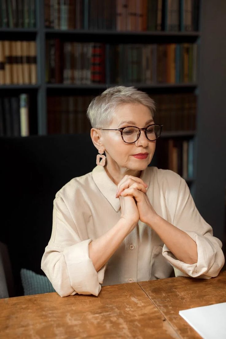 an older woman sitting at a table in front of bookshelves with her hands folded