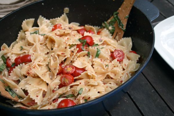 a skillet filled with pasta and tomatoes on top of a wooden table next to a white plate