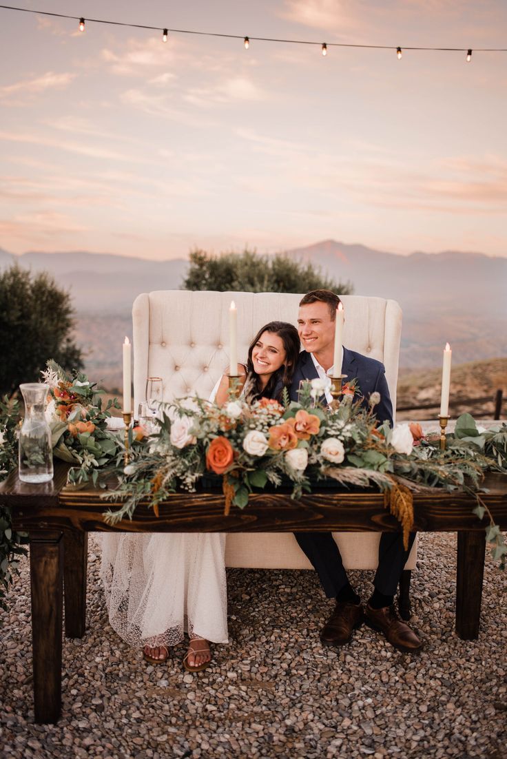 a newly married couple sitting on a bench with flowers and candles in front of them