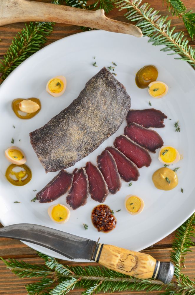 a white plate topped with meat and olives next to a knife on top of a wooden table