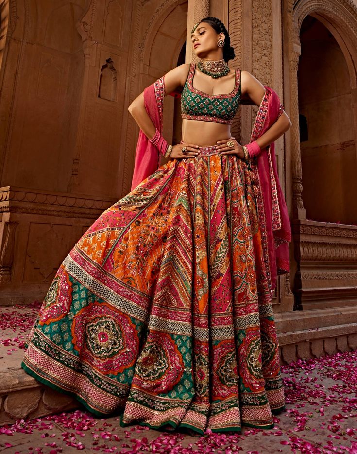 Aadhya lehenga set is an eye-catching piece crafted in multi-coloured festive colours printed silk, featuring heavy tassel strings with gota embroidery, hand adda work, and beads/sequins. The bold colours of pink, green and orange add a vibrant touch to this one-of-a-kind set. Orange Lengha Color Combos, Multi Color Mehendi Outfit, Multicolor Lehenga Bridal, Sabyasachi Multicolor Lehenga, Printed Indian Outfits, Semi Traditional Outfits For Women, Multi Colour Lehenga Indian Weddings, Traditional Nepali Clothing, Funky Lehenga