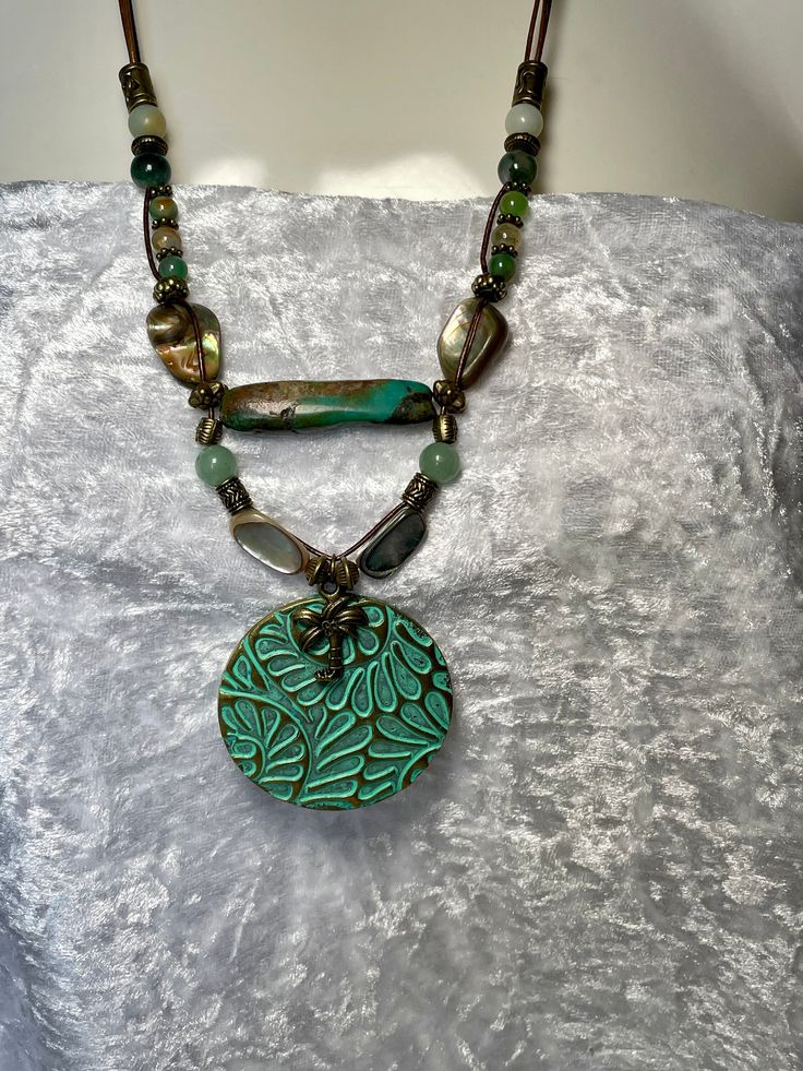 Fun, bold colorful 2" pendant with Palm Tree charm on two strands of 1mm bronze leather cord, Peacock Agate, Aventurine, India Agate and two 3/4" each Mother of Pearl. A large Genuine 1 1/2" Sea Green Turquoise Bead takes center stage bringing the colors together. Bronze toggle to close. Necklace is 22"  around neck with drop of pendant  total is 24". No lead or nickel used. Please use the Last On, First Off approach. Stones discolor with lotions, perfumes, hairspray, sweat, chlorine, even some soaps, and will break if dropped. Green Necklace With Large Medallion Pendant, Green Beaded Necklace With Round Pendant, Green Bohemian Double Strand Jewelry, Bohemian Green Pendant Beaded Necklace, Bohemian Green Beaded Pendant Necklace, Green Double Strand Spiritual Necklace, Artisan Green Turquoise Necklace With Large Pendant, Unique Turquoise Double Strand Necklace, Handmade Green Turquoise Bohemian Necklace