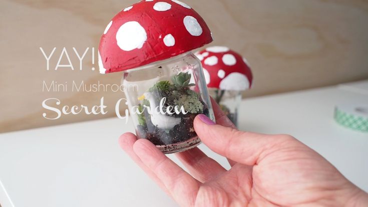 a hand holding a small jar filled with dirt and plants next to a white table