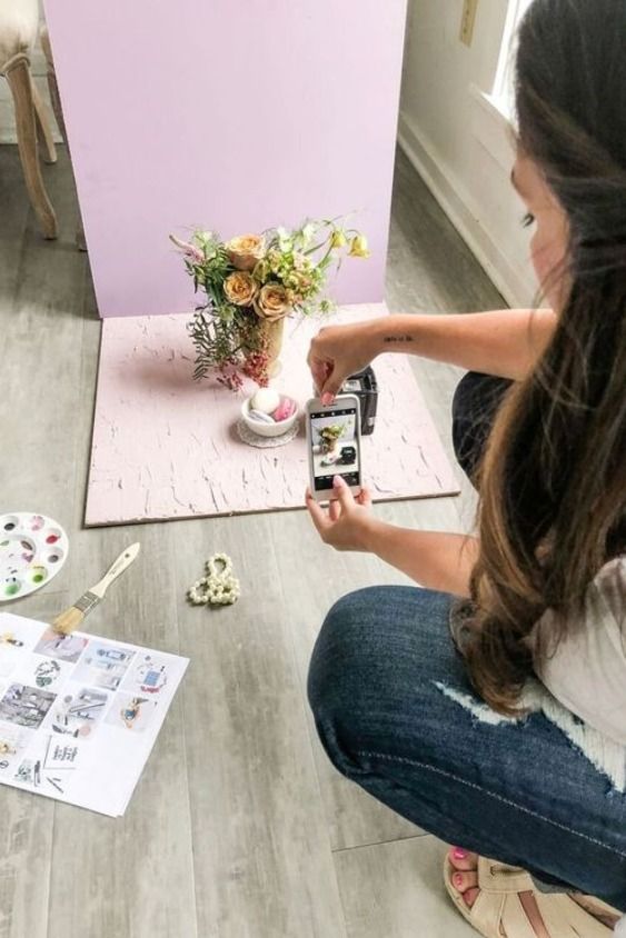 a woman sitting on the floor taking pictures with her cell phone and painting flowers in a vase