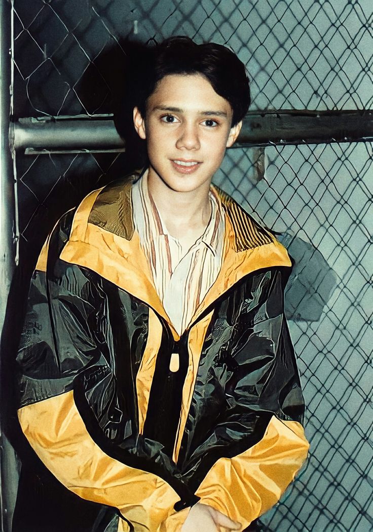 a young man in a yellow and black jacket standing next to a chain link fence