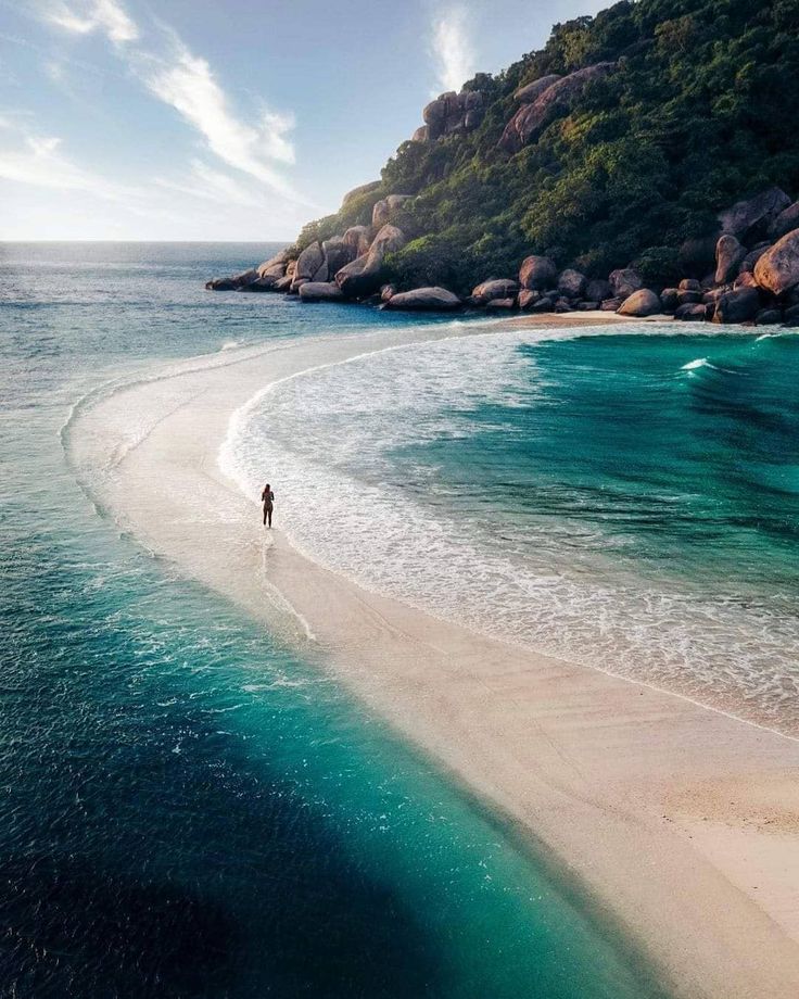 a person is walking along the beach in front of some rocks and water with an island in the background