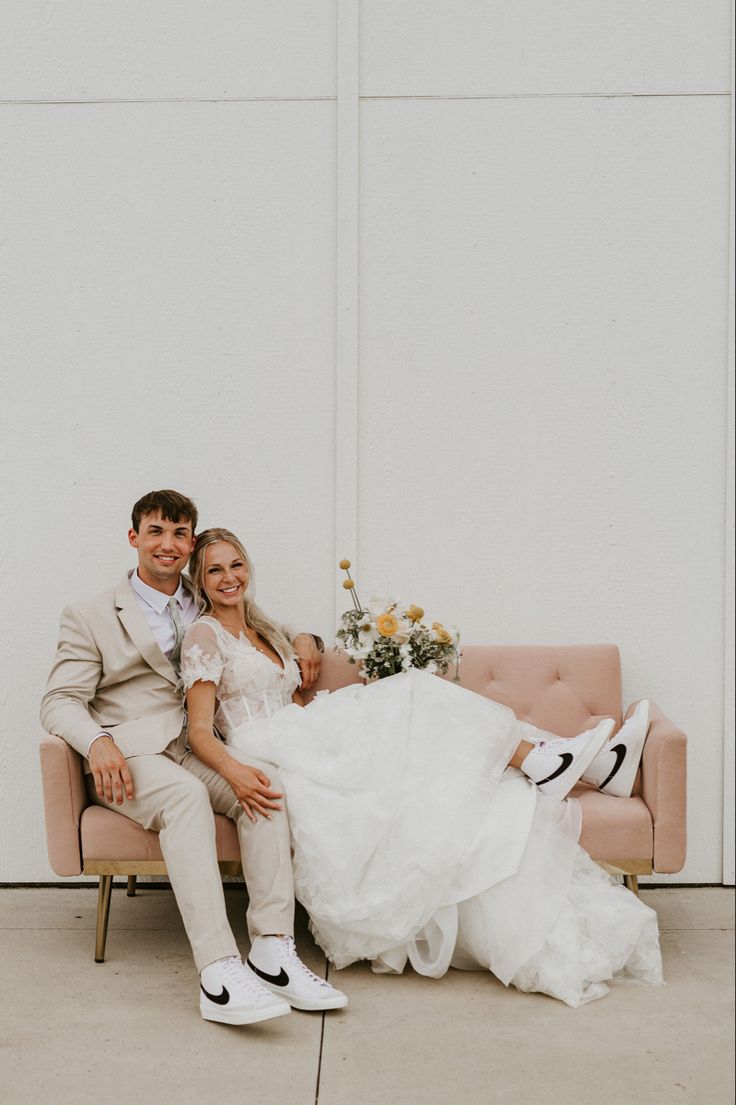 a bride and groom sitting on a couch in front of a white wall with flowers