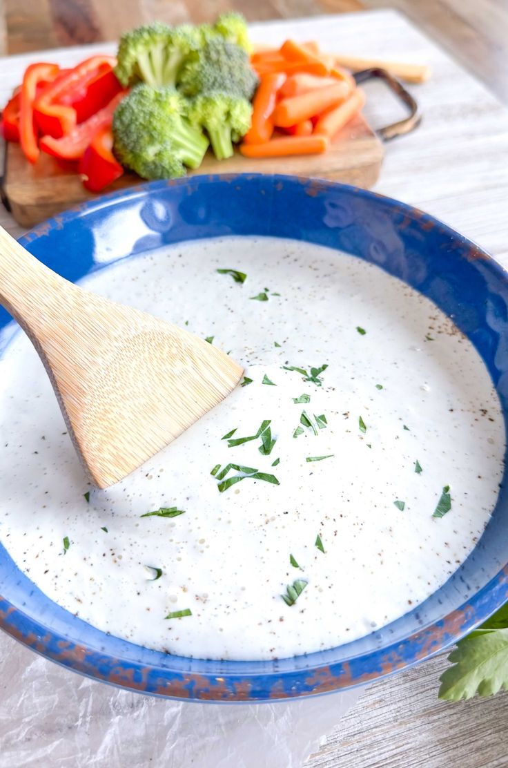 a wooden spoon in a blue bowl filled with white sauce next to broccoli and carrots