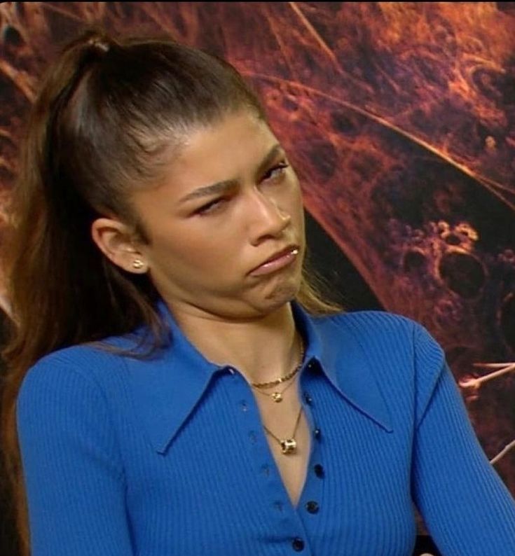 a woman in a blue shirt sitting down with her hand on her hip and looking at the camera