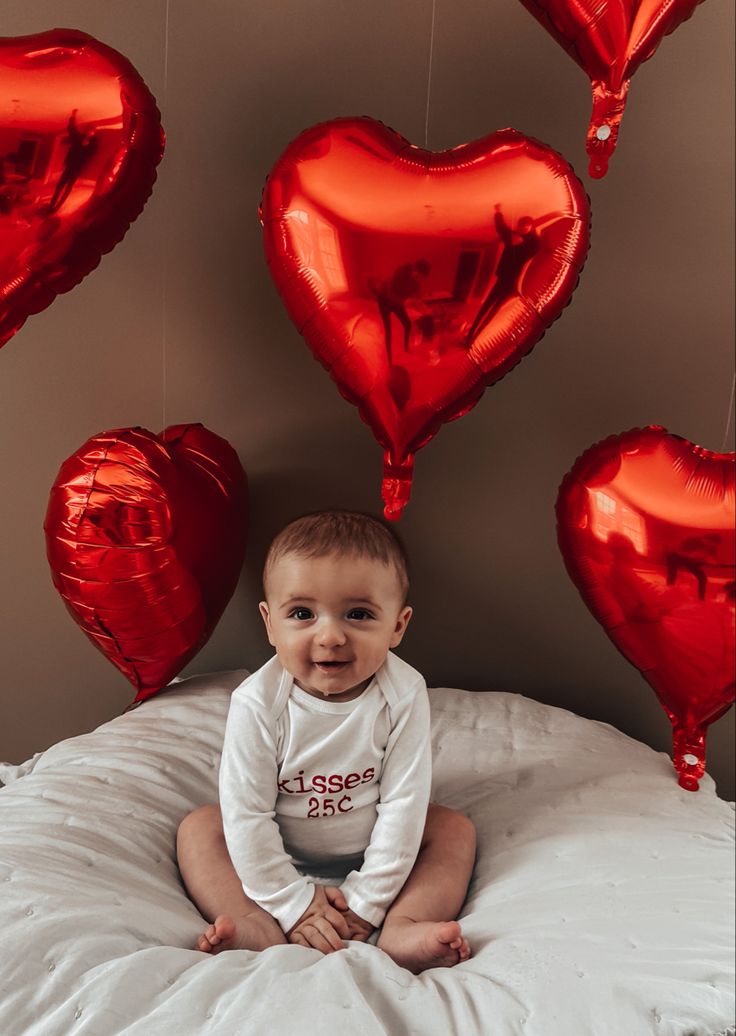 a baby sitting on a bed surrounded by red heart balloons