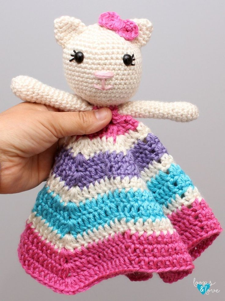 a hand holding a small crocheted cat in a colorful dress with pink, blue and white stripes