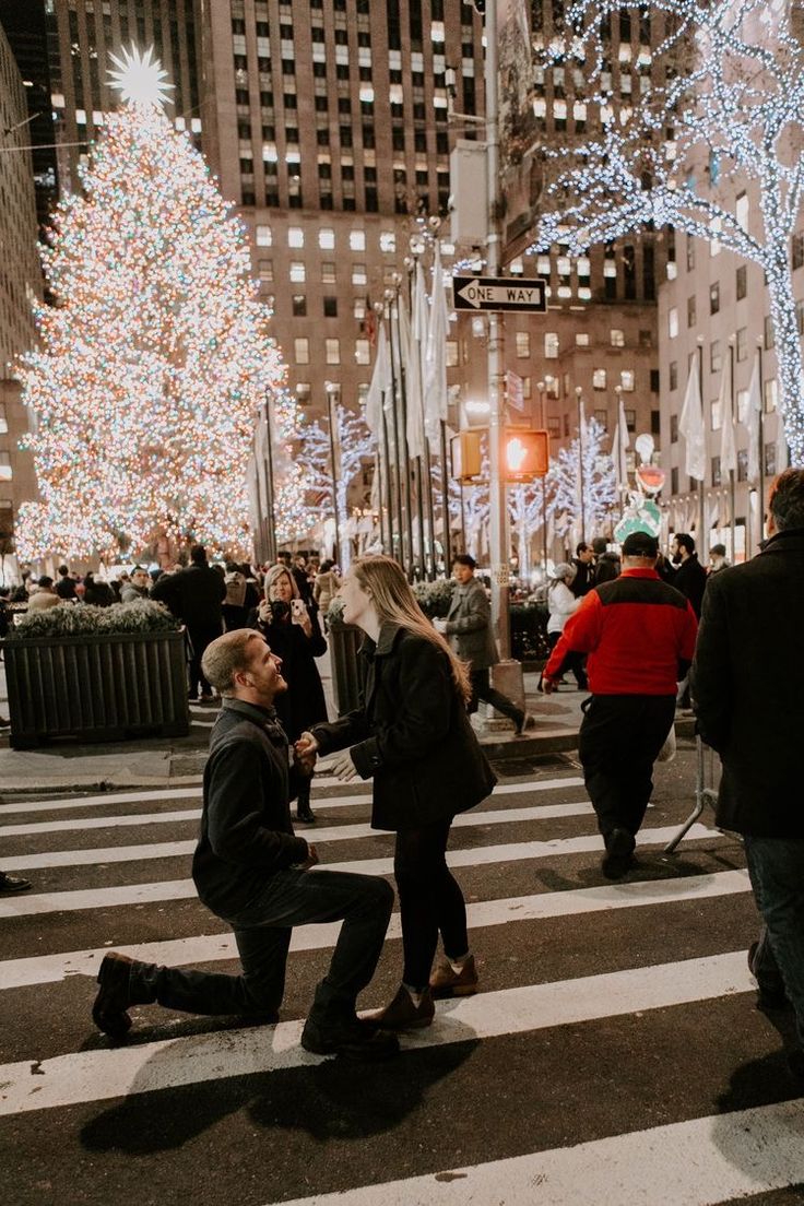 two people kneeling down in the middle of a cross walk at night with christmas lights all around them