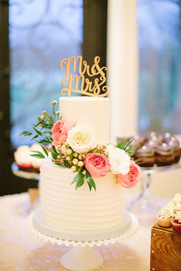 a white wedding cake with pink flowers and mr and mrs sign on top, sitting on a table in front of cupcakes