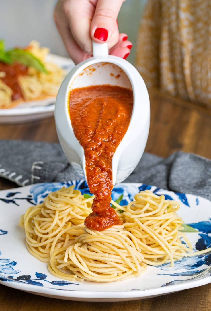 a woman pouring sauce over spaghetti on top of a blue and white plate with another plate in the background