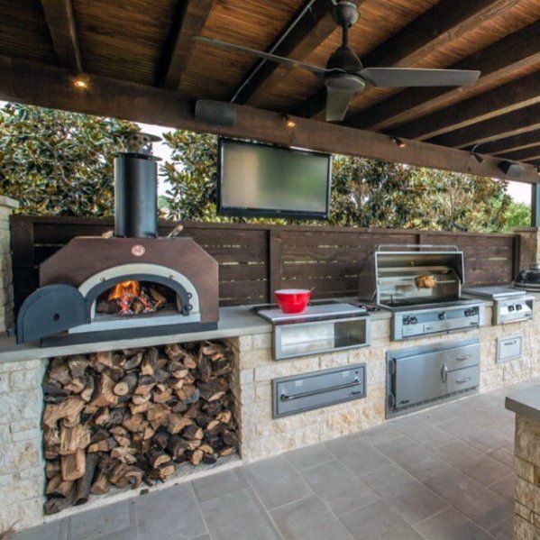 an outdoor kitchen with pizza oven, grill and television on the outside wall above it