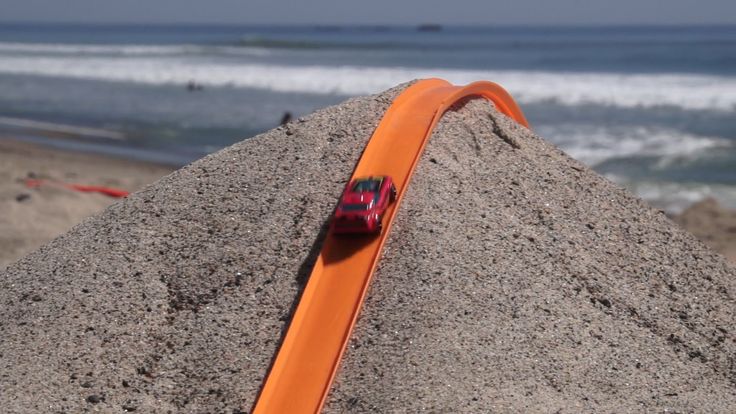 an orange toy car is on top of a sand dune at the beach with waves in the background