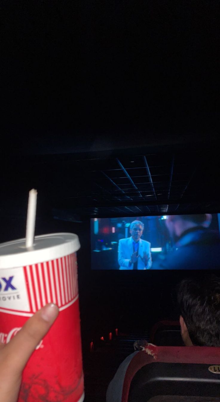 a person holding up a cup with a straw in it while watching a movie on the screen