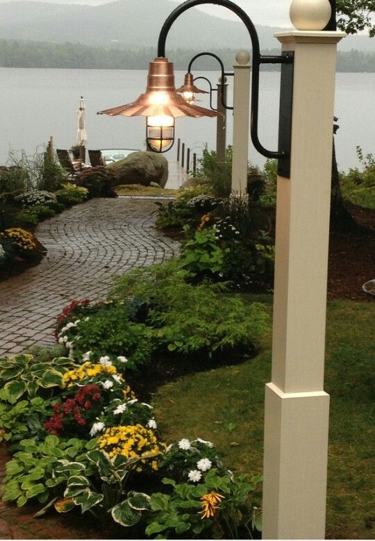 a light that is on the side of a pole near some flowers and plants in front of a body of water