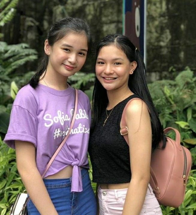 two young women standing next to each other in front of trees and bushes, one holding a pink handbag