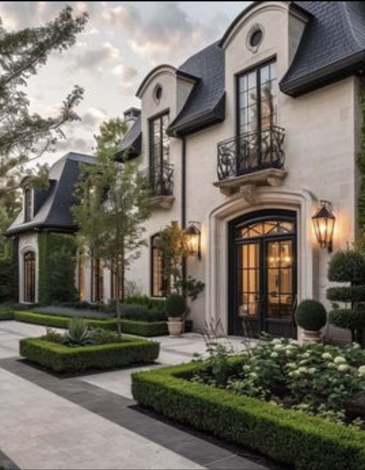 a large white house surrounded by hedges and trees with lights on the front door, along with potted plants