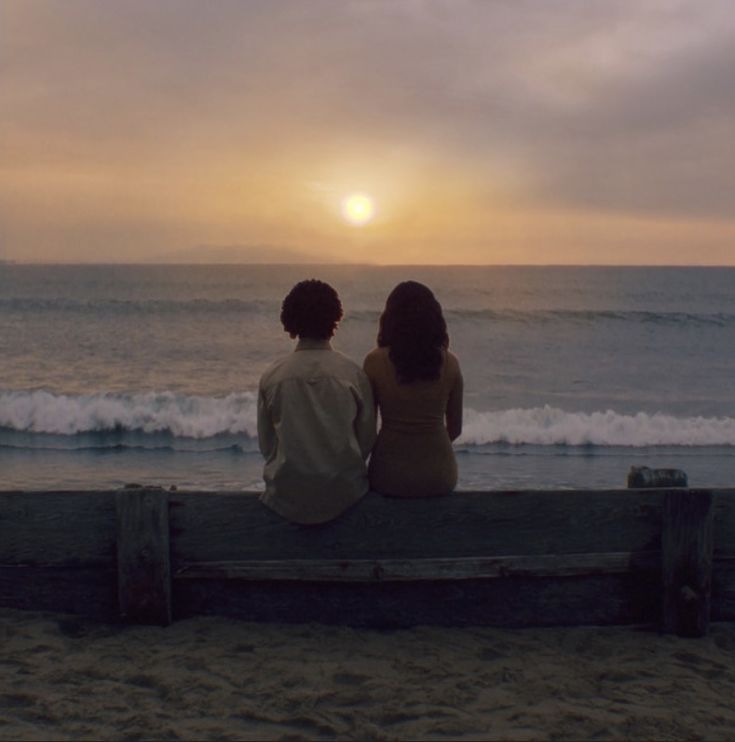 two people sitting on a bench looking out at the ocean while the sun is setting