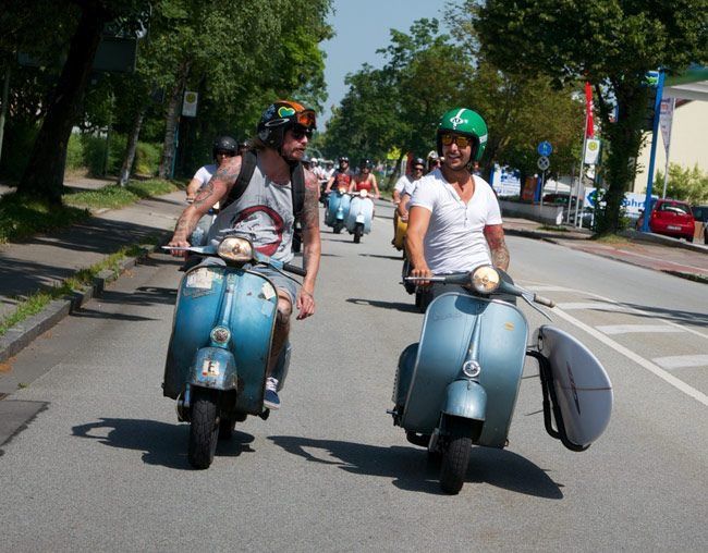 two men riding on the back of scooters down a street with trees in the background