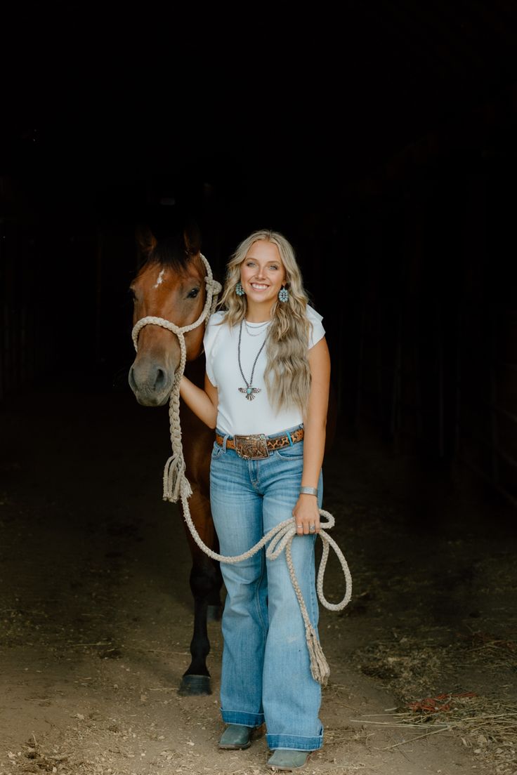 a woman standing next to a brown horse in a dark barn with a rope around her neck