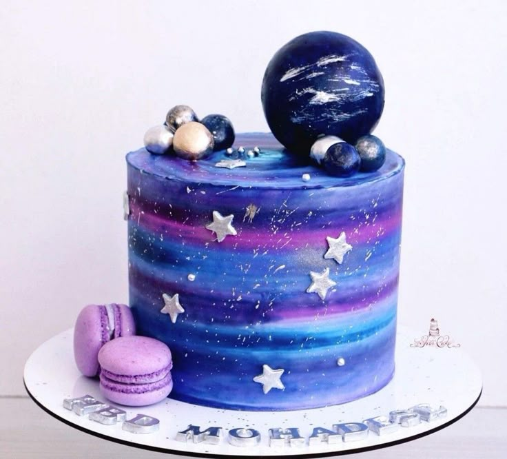 a cake decorated with stars and planets on top of a white plate, next to two macaroons