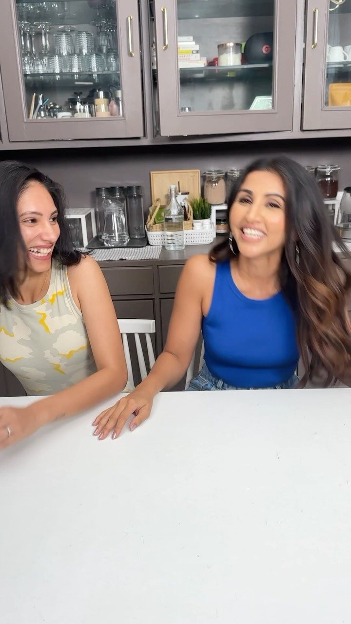 two women sitting at a table in a kitchen smiling and looking into the camera,