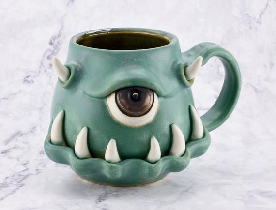 a green ceramic mug with an evil looking monster's head on it, sitting on a marble surface