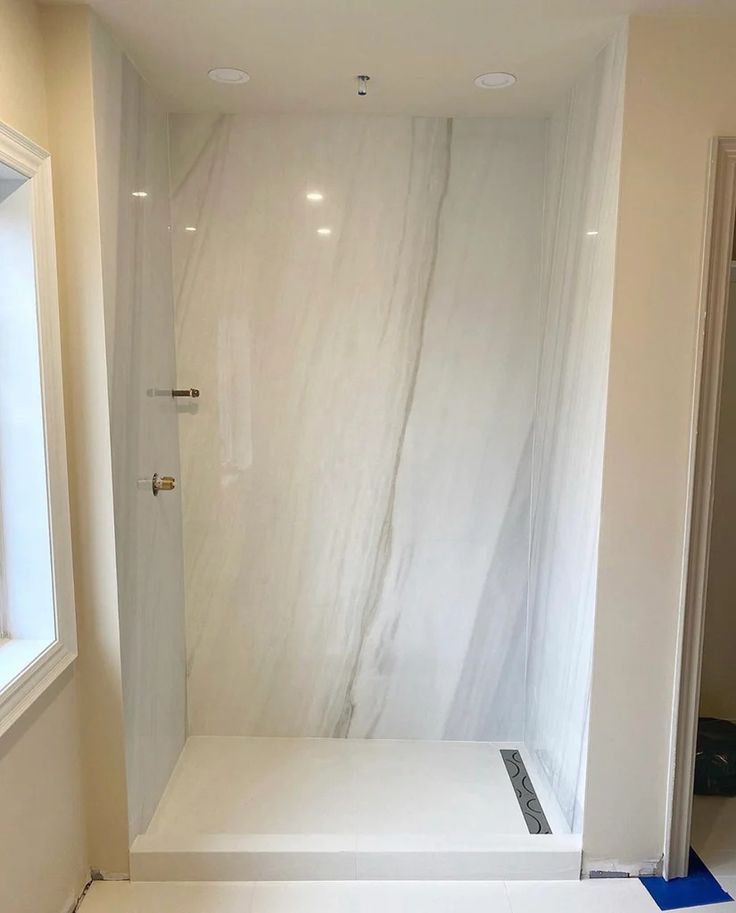 an empty bathroom with white marble walls and flooring in the shower area, next to a window