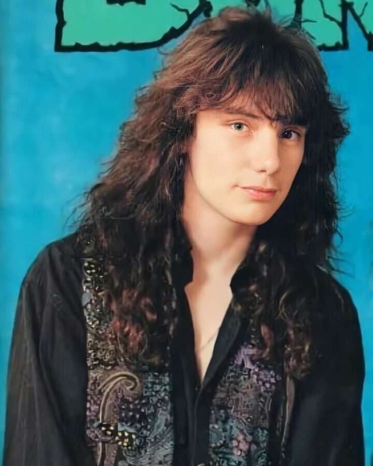 a young man with long hair wearing a black shirt and looking at the camera while standing in front of a blue background