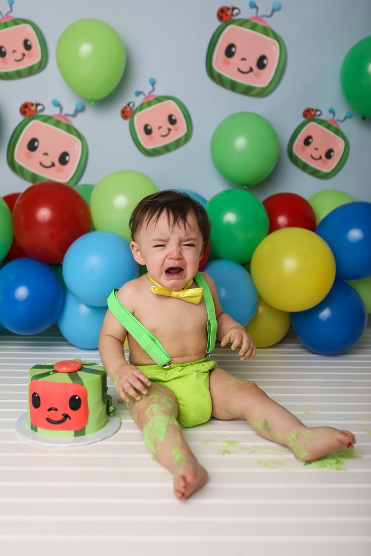 a baby sitting on the floor in front of balloons and cake with his mouth open