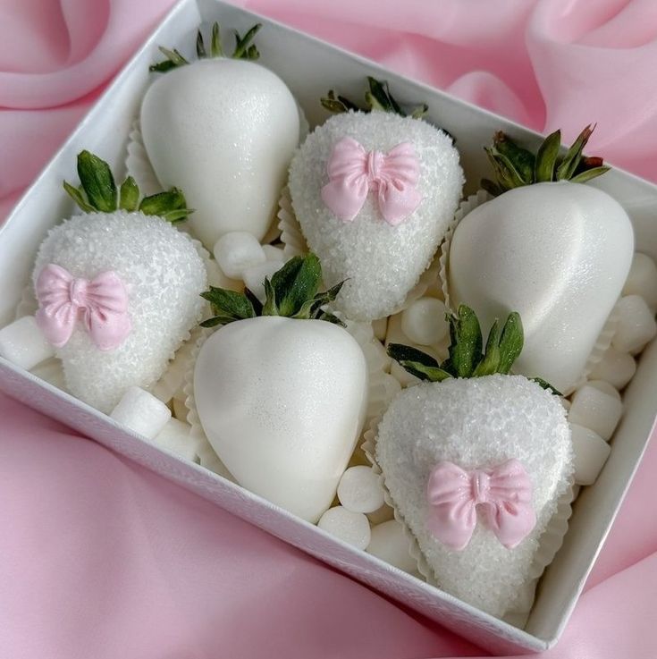 four white chocolate covered strawberries with pink bows in a square box on a pink cloth