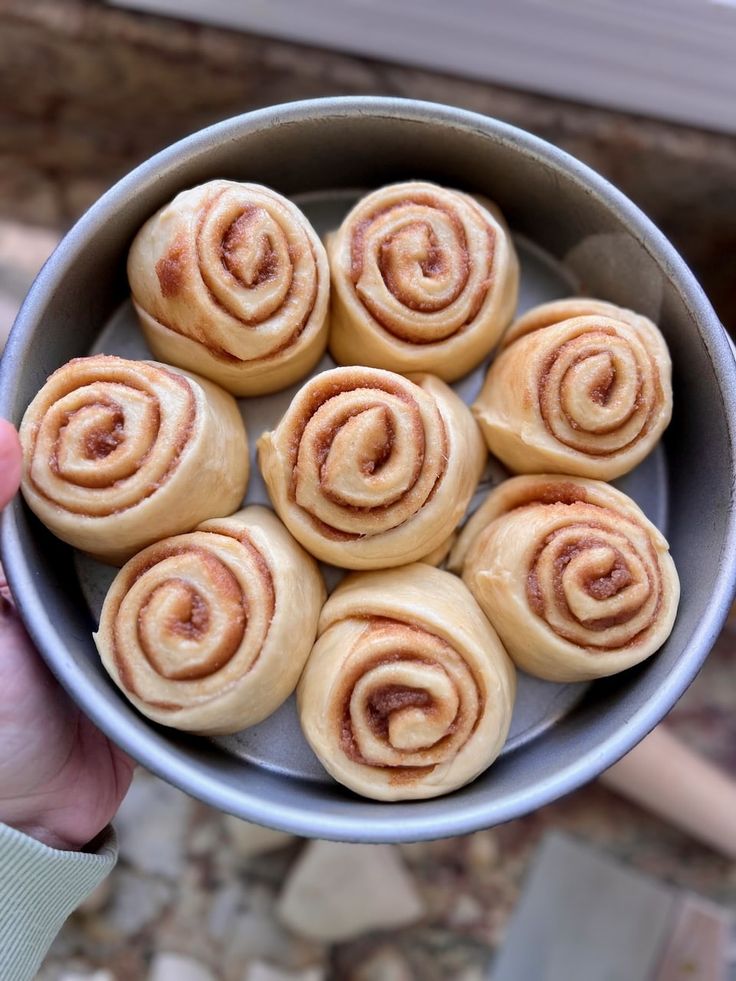 a person holding a metal bowl filled with cinnamon buns on top of a table