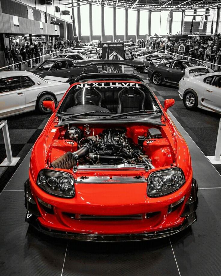 an orange sports car with its hood open in a garage filled with other cars and people