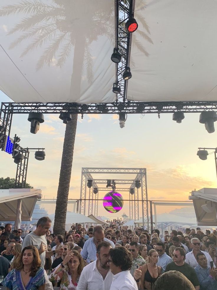 a large group of people sitting under a canopy at an outdoor music festival with the sun setting in the background