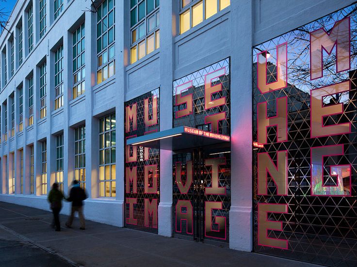 two people are walking down the sidewalk in front of a building that has neon letters on it