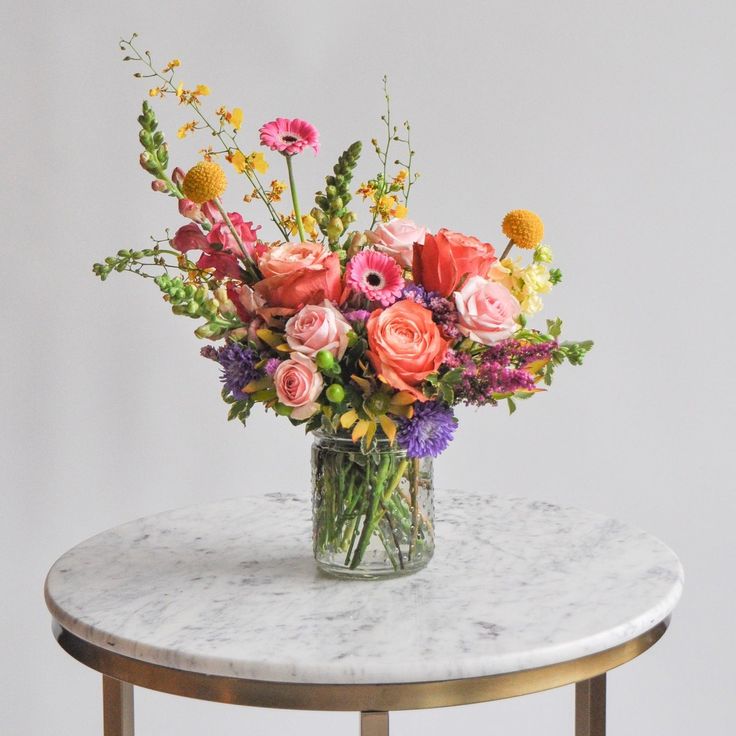 A glass vase with a medium flower arrangement sits on a marble table in front of a white backdrop. The flowers are many colors and include roses Flower Arrangements For Table Birthday, Artificial Wildflower Arrangements, Wildflower Bouquet In Vase, Live Flower Wedding Bouquets, Everyday Floral Arrangements, Asymmetrical Flower Arrangement, Summer Wedding Flowers Bouquets, Bohemian Flower Arrangements, Wildflower Centerpiece Wedding