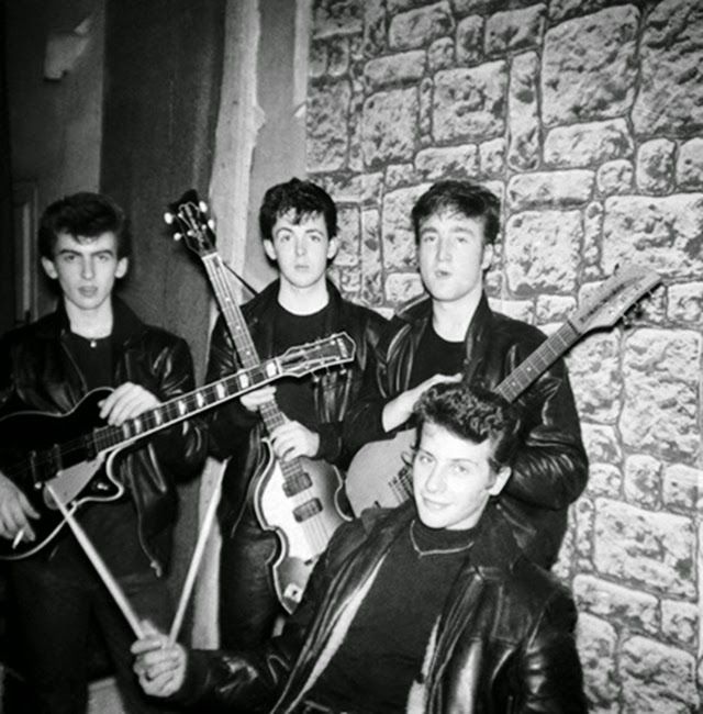 four young men are posing with their instruments in front of a brick wall, one is wearing a black leather jacket and the other has a guitar