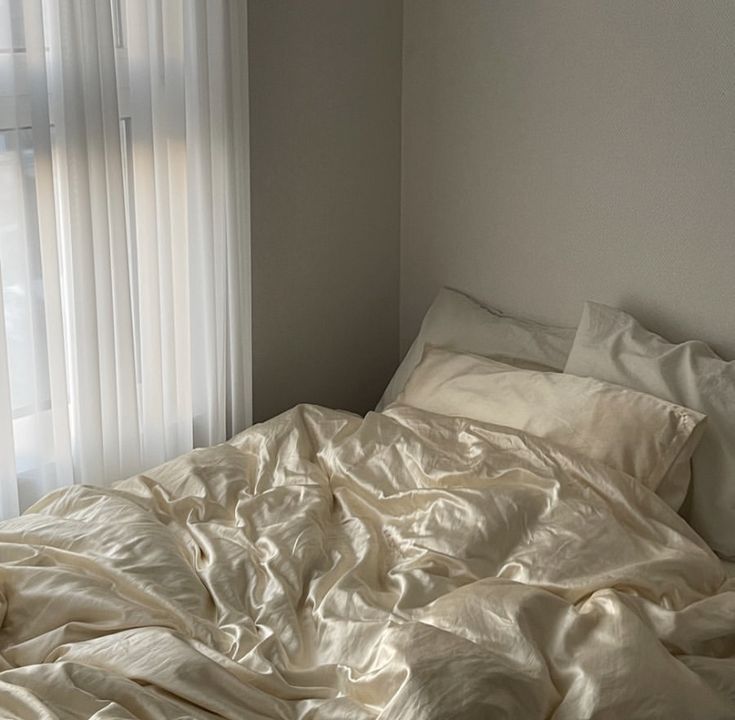 an unmade bed in front of a window with white sheets and pillows on it