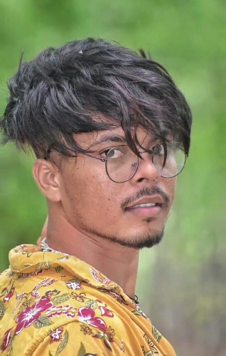 a man with glasses and a yellow shirt looks at the camera while standing in front of some trees