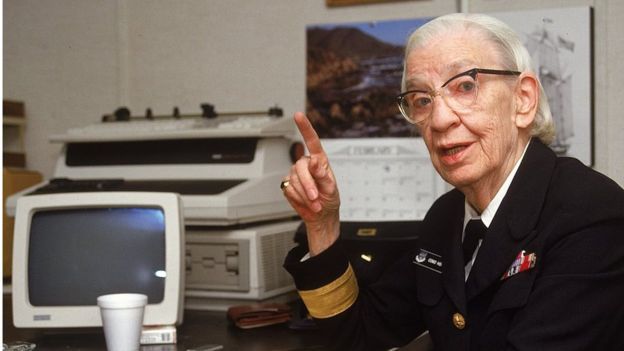 an older woman in uniform sitting at a desk pointing to the side with her finger