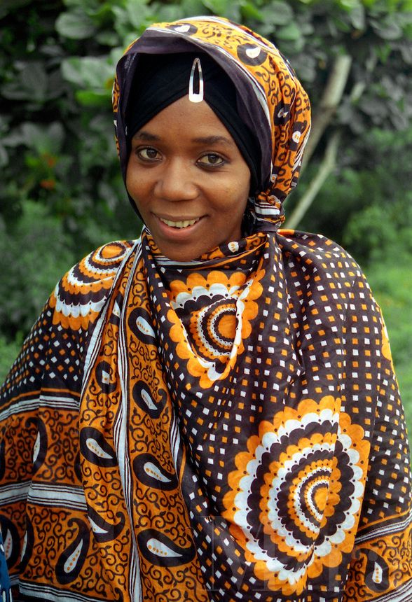 a woman in an orange and black patterned shawl smiles at the camera with trees behind her