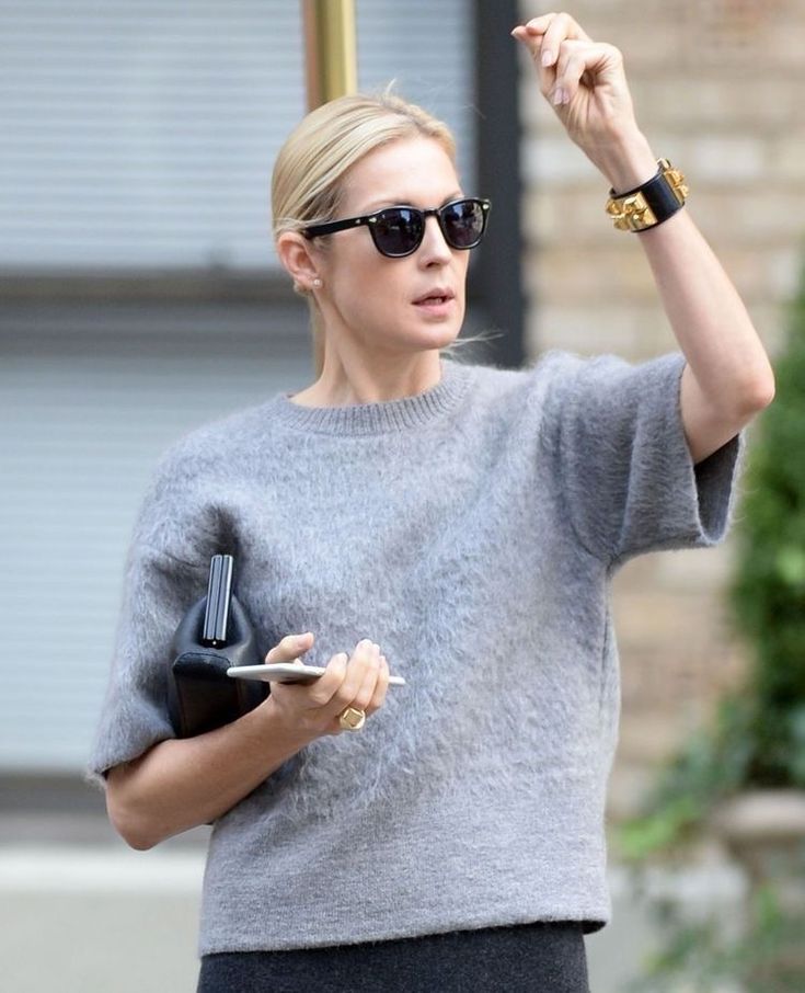 a woman in grey sweater and black skirt pointing at something on her hand while holding a cell phone