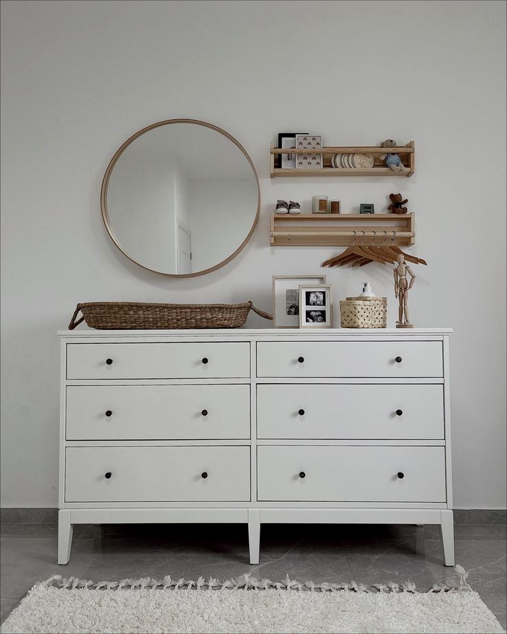 a white dresser with drawers and a round mirror on the wall above it, next to a shelf filled with books