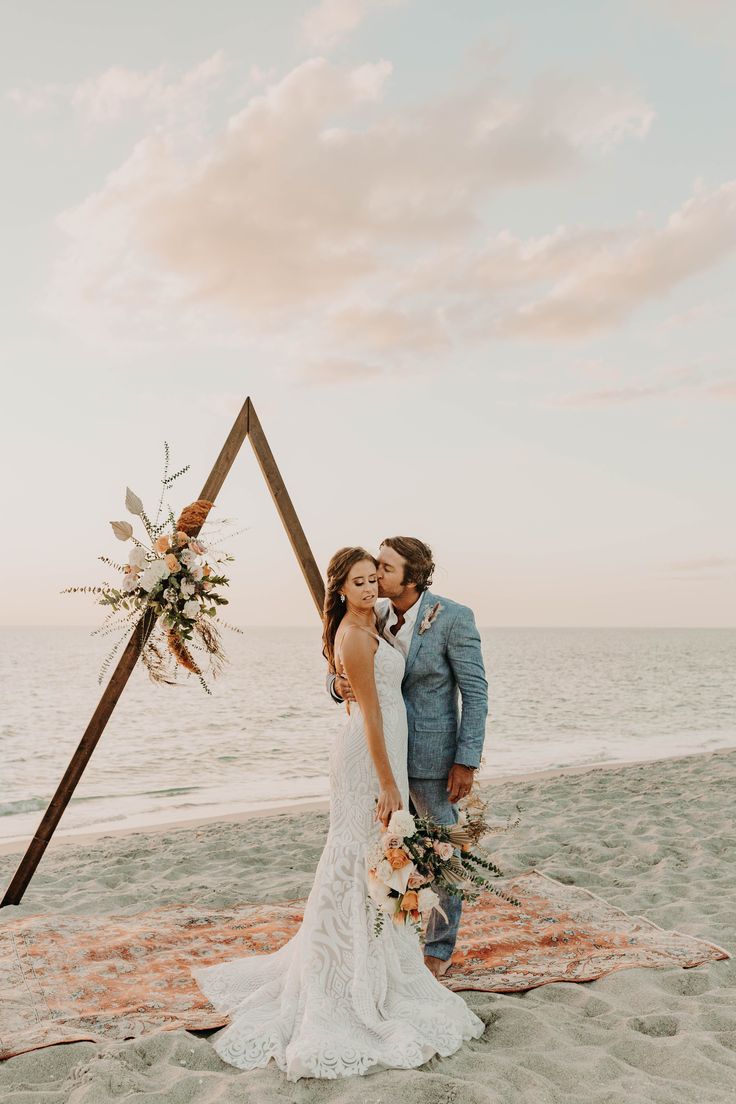 a bride and groom kissing on the beach in front of an arch decorated with flowers