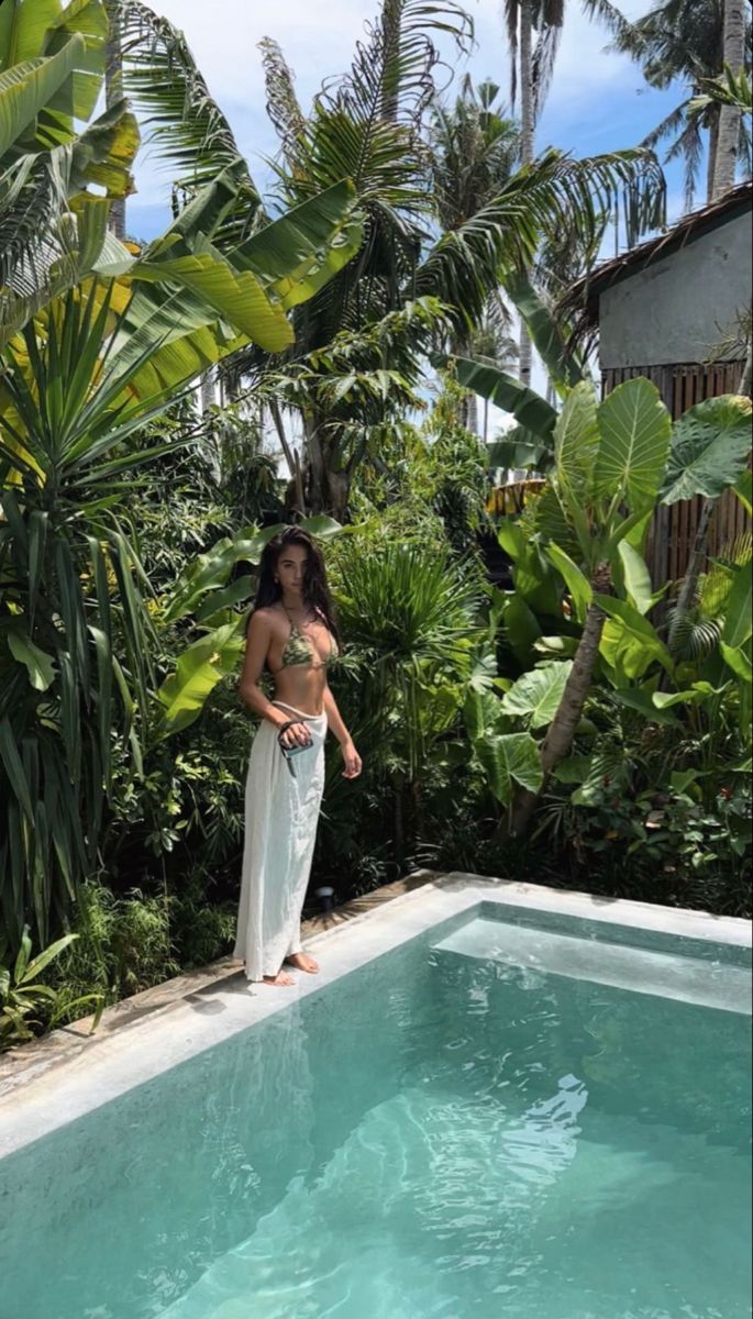 a woman standing in front of a swimming pool surrounded by palm trees and greenery