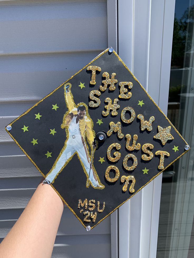 a hand holding a graduation cap that says the show must go on with gold stars